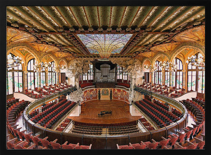A high-angle, wide shot of the ornate and lavish interior of the Palau de la Música Catalana in Barcelona. The view captures the entire concert hall, with its curved balconies filled with red seats, a central wooden stage with a grand piano, and a spectacular, large stained-glass skylight in the ceiling. Poster