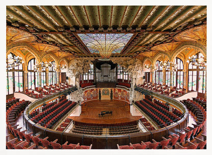 A high-angle, wide shot of the ornate and lavish interior of the Palau de la Música Catalana in Barcelona. The view captures the entire concert hall, with its curved balconies filled with red seats, a central wooden stage with a grand piano, and a spectacular, large stained-glass skylight in the ceiling. Poster