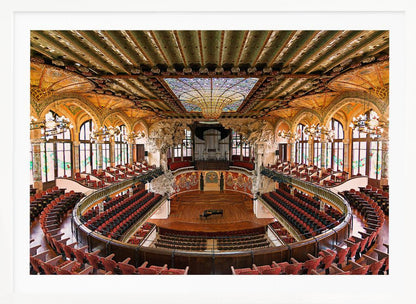 A high-angle, wide shot of the ornate and lavish interior of the Palau de la Música Catalana in Barcelona. The view captures the entire concert hall, with its curved balconies filled with red seats, a central wooden stage with a grand piano, and a spectacular, large stained-glass skylight in the ceiling. Poster