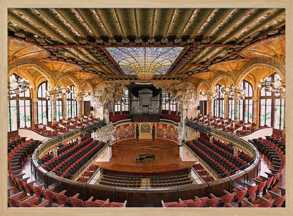 A high-angle, wide shot of the ornate and lavish interior of the Palau de la Música Catalana in Barcelona. The view captures the entire concert hall, with its curved balconies filled with red seats, a central wooden stage with a grand piano, and a spectacular, large stained-glass skylight in the ceiling. Poster