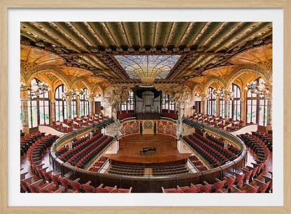 A high-angle, wide shot of the ornate and lavish interior of the Palau de la Música Catalana in Barcelona. The view captures the entire concert hall, with its curved balconies filled with red seats, a central wooden stage with a grand piano, and a spectacular, large stained-glass skylight in the ceiling. Poster