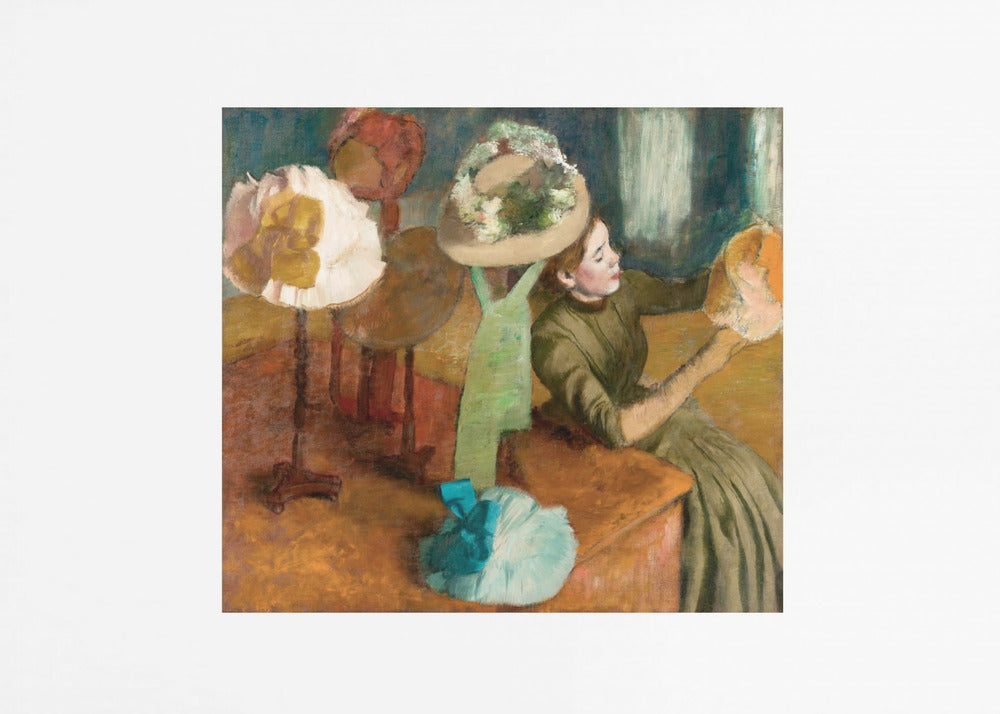 A framed print of Edgar Degas's Impressionist painting 'The Millinery Shop,' showing a woman in a green dress examining a hat in a shop filled with various fashionable hats on display stands. Print