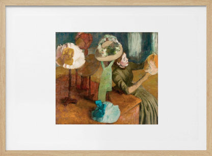 A framed print of Edgar Degas's Impressionist painting 'The Millinery Shop,' showing a woman in a green dress examining a hat in a shop filled with various fashionable hats on display stands. Print