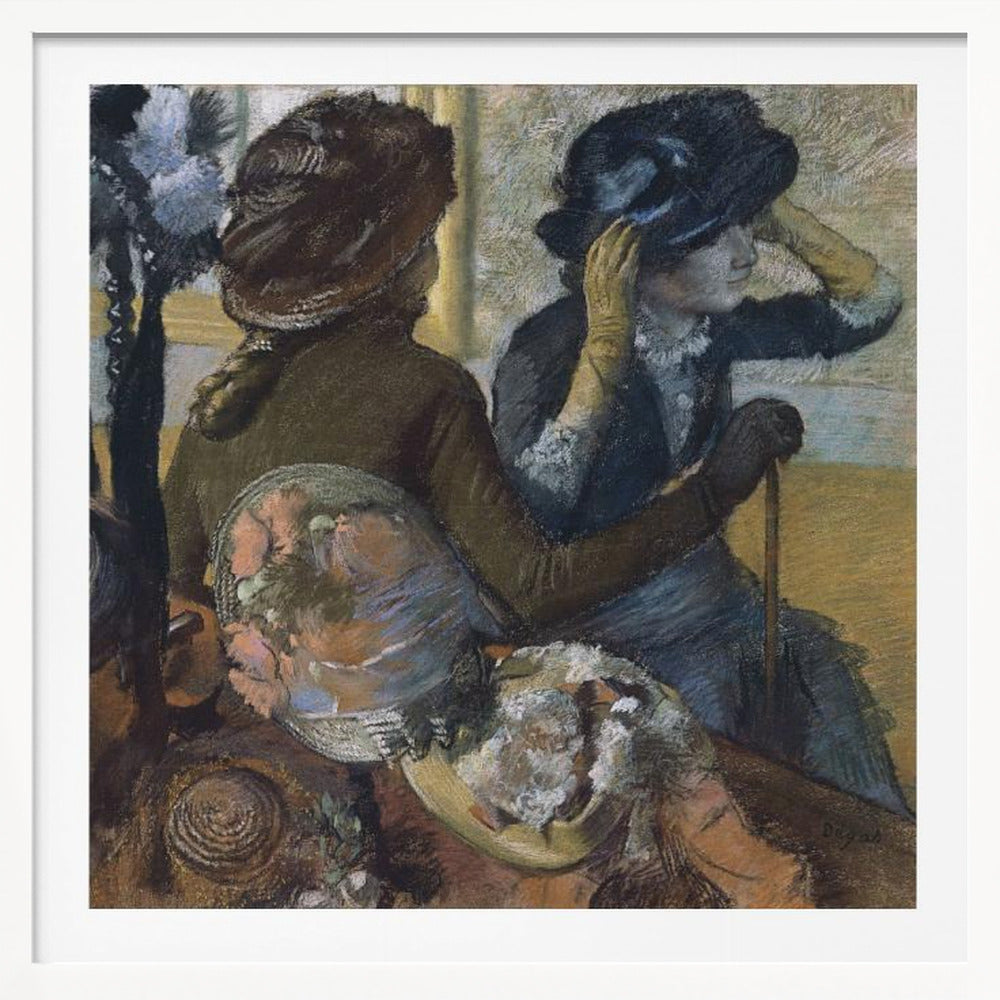 A framed impressionist painting by Edgar Degas, 'The Millinery Shop', depicting two women. One woman, seen from the back, wears a brown coat and hat, while the other tries on a dark blue hat, adjusting it with her gloved hands. Colorful hats are displayed in the foreground. Print
