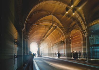 A long exposure photograph of a grand, vaulted hallway with brick arches. Dramatic beams of golden light cut through the space, creating horizontal streaks and a motion blur effect. Silhouettes of people walk through the atmospheric light and shadow. Wall Art