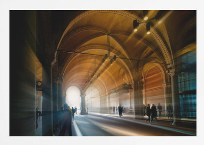 A long exposure photograph of a grand, vaulted hallway with brick arches. Dramatic beams of golden light cut through the space, creating horizontal streaks and a motion blur effect. Silhouettes of people walk through the atmospheric light and shadow. Wall Art