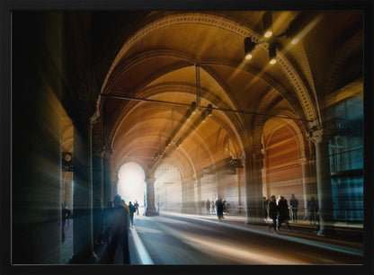 A long exposure photograph of a grand, vaulted hallway with brick arches. Dramatic beams of golden light cut through the space, creating horizontal streaks and a motion blur effect. Silhouettes of people walk through the atmospheric light and shadow. Wall Art