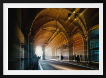 A long exposure photograph of a grand, vaulted hallway with brick arches. Dramatic beams of golden light cut through the space, creating horizontal streaks and a motion blur effect. Silhouettes of people walk through the atmospheric light and shadow. Wall Art