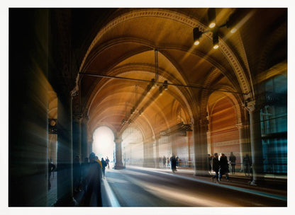 A long exposure photograph of a grand, vaulted hallway with brick arches. Dramatic beams of golden light cut through the space, creating horizontal streaks and a motion blur effect. Silhouettes of people walk through the atmospheric light and shadow. Wall Art