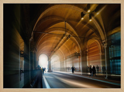 A long exposure photograph of a grand, vaulted hallway with brick arches. Dramatic beams of golden light cut through the space, creating horizontal streaks and a motion blur effect. Silhouettes of people walk through the atmospheric light and shadow. Wall Art