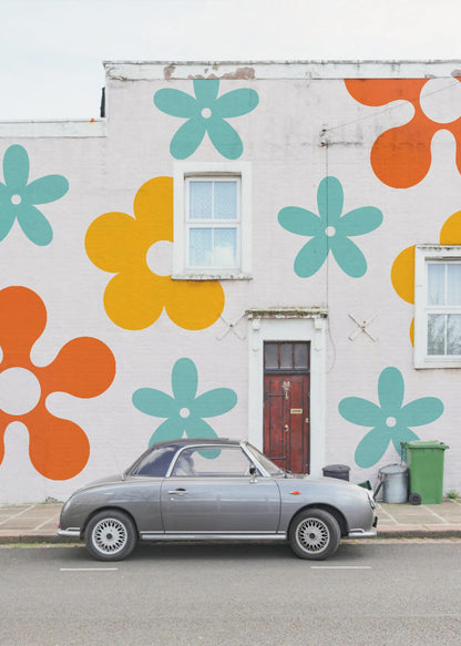 A retro-style gray convertible parked on a city street in front of a building with a large, colorful pop-art mural of stylized flowers in orange, yellow, and turquoise. Decor