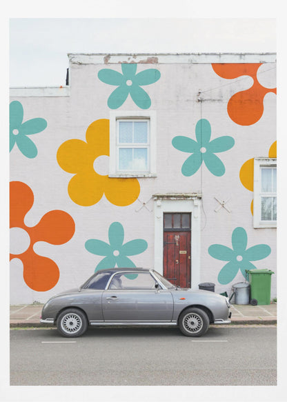 A retro-style gray convertible parked on a city street in front of a building with a large, colorful pop-art mural of stylized flowers in orange, yellow, and turquoise. Decor