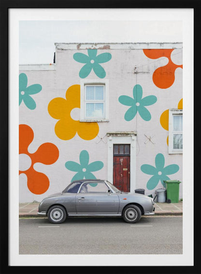 A retro-style gray convertible parked on a city street in front of a building with a large, colorful pop-art mural of stylized flowers in orange, yellow, and turquoise. Decor