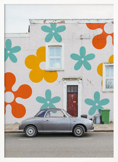 A retro-style gray convertible parked on a city street in front of a building with a large, colorful pop-art mural of stylized flowers in orange, yellow, and turquoise. Decor