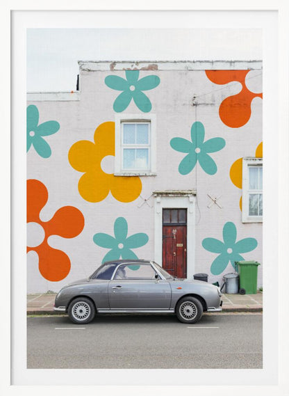 A retro-style gray convertible parked on a city street in front of a building with a large, colorful pop-art mural of stylized flowers in orange, yellow, and turquoise. Decor