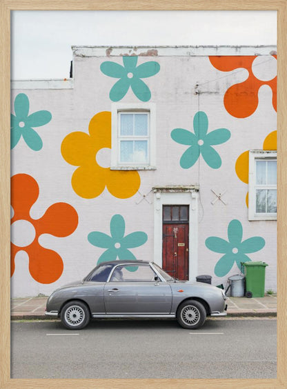 A retro-style gray convertible parked on a city street in front of a building with a large, colorful pop-art mural of stylized flowers in orange, yellow, and turquoise. Decor