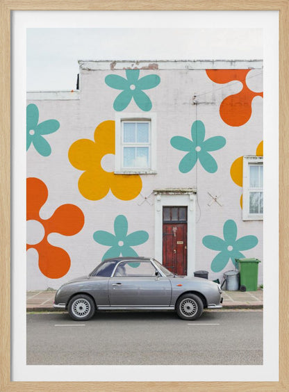 A retro-style gray convertible parked on a city street in front of a building with a large, colorful pop-art mural of stylized flowers in orange, yellow, and turquoise. Decor