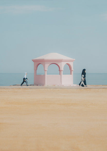 A minimalist photograph of a coastal scene with a pastel color palette. A pink arched gazebo sits on a promenade with the blue sea and sky in the background. A large expanse of beige sand is in the foreground. Two figures in hijabs walk in opposite directions on either side of the gazebo, one with a small child. Decor