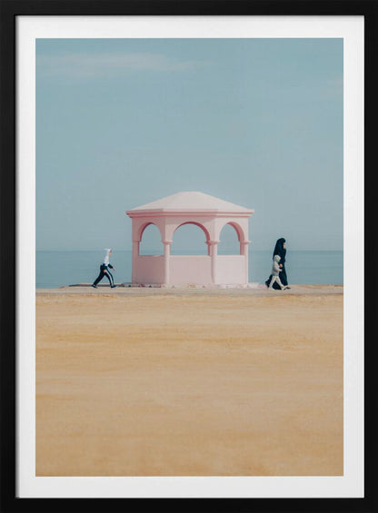 A minimalist photograph of a coastal scene with a pastel color palette. A pink arched gazebo sits on a promenade with the blue sea and sky in the background. A large expanse of beige sand is in the foreground. Two figures in hijabs walk in opposite directions on either side of the gazebo, one with a small child. Decor