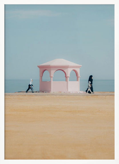 A minimalist photograph of a coastal scene with a pastel color palette. A pink arched gazebo sits on a promenade with the blue sea and sky in the background. A large expanse of beige sand is in the foreground. Two figures in hijabs walk in opposite directions on either side of the gazebo, one with a small child. Decor