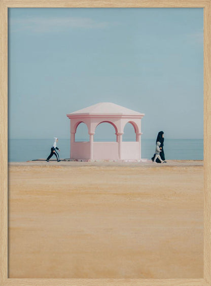A minimalist photograph of a coastal scene with a pastel color palette. A pink arched gazebo sits on a promenade with the blue sea and sky in the background. A large expanse of beige sand is in the foreground. Two figures in hijabs walk in opposite directions on either side of the gazebo, one with a small child. Decor