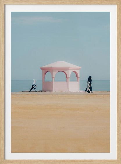 A minimalist photograph of a coastal scene with a pastel color palette. A pink arched gazebo sits on a promenade with the blue sea and sky in the background. A large expanse of beige sand is in the foreground. Two figures in hijabs walk in opposite directions on either side of the gazebo, one with a small child. Decor