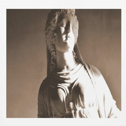 A framed, sepia-toned photograph of a classical marble bust of a woman. She looks upwards and to the side, illuminated by a strong light that casts dramatic shadows across her face and draped clothing. The photo is enclosed in a simple, light wood frame. Print