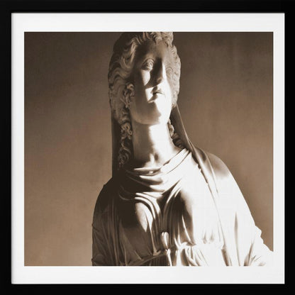 A framed, sepia-toned photograph of a classical marble bust of a woman. She looks upwards and to the side, illuminated by a strong light that casts dramatic shadows across her face and draped clothing. The photo is enclosed in a simple, light wood frame. Print