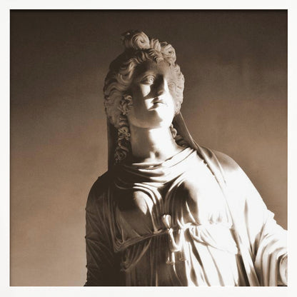 A framed, sepia-toned photograph of a classical marble bust of a woman. She looks upwards and to the side, illuminated by a strong light that casts dramatic shadows across her face and draped clothing. The photo is enclosed in a simple, light wood frame. Print