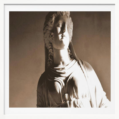 A framed, sepia-toned photograph of a classical marble bust of a woman. She looks upwards and to the side, illuminated by a strong light that casts dramatic shadows across her face and draped clothing. The photo is enclosed in a simple, light wood frame. Print
