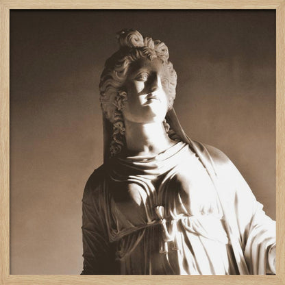 A framed, sepia-toned photograph of a classical marble bust of a woman. She looks upwards and to the side, illuminated by a strong light that casts dramatic shadows across her face and draped clothing. The photo is enclosed in a simple, light wood frame. Print
