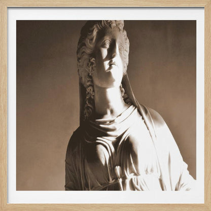 A framed, sepia-toned photograph of a classical marble bust of a woman. She looks upwards and to the side, illuminated by a strong light that casts dramatic shadows across her face and draped clothing. The photo is enclosed in a simple, light wood frame. Print