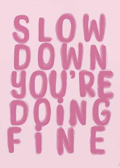 A framed art print featuring the phrase 'SLOW DOWN YOU'RE DOING FINE' in a bold, dark pink, brush-stroke font. The words are stacked vertically against a light pink background, all enclosed within a simple black frame. Artwork