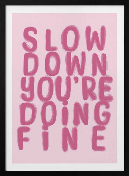 A framed art print featuring the phrase 'SLOW DOWN YOU'RE DOING FINE' in a bold, dark pink, brush-stroke font. The words are stacked vertically against a light pink background, all enclosed within a simple black frame. Artwork