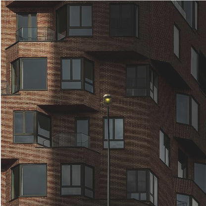 A close-up photograph of a modern red-brick apartment building, framed in light wood. A sharp shadow line divides the facade, with a lit streetlamp providing a warm yellow glow in the shaded area. Decor