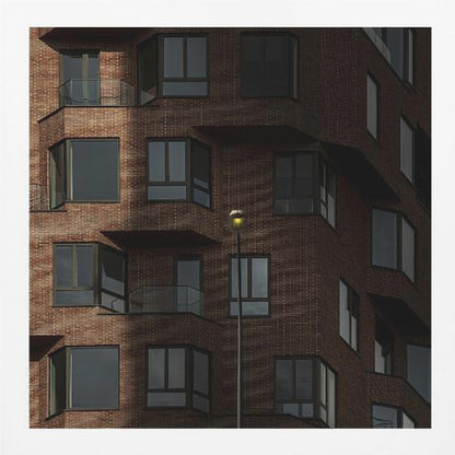 A close-up photograph of a modern red-brick apartment building, framed in light wood. A sharp shadow line divides the facade, with a lit streetlamp providing a warm yellow glow in the shaded area. Decor