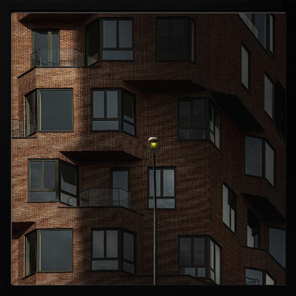 A close-up photograph of a modern red-brick apartment building, framed in light wood. A sharp shadow line divides the facade, with a lit streetlamp providing a warm yellow glow in the shaded area. Decor