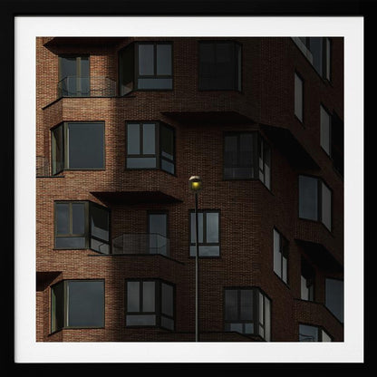 A close-up photograph of a modern red-brick apartment building, framed in light wood. A sharp shadow line divides the facade, with a lit streetlamp providing a warm yellow glow in the shaded area. Decor