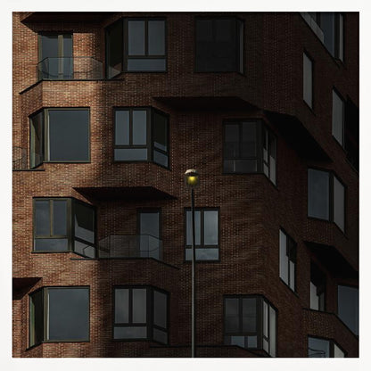 A close-up photograph of a modern red-brick apartment building, framed in light wood. A sharp shadow line divides the facade, with a lit streetlamp providing a warm yellow glow in the shaded area. Decor