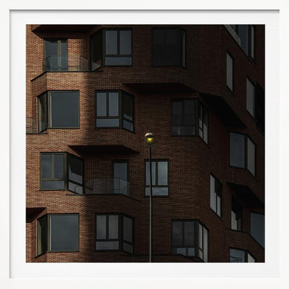A close-up photograph of a modern red-brick apartment building, framed in light wood. A sharp shadow line divides the facade, with a lit streetlamp providing a warm yellow glow in the shaded area. Decor