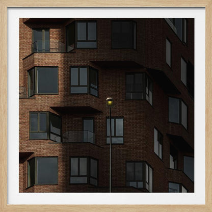 A close-up photograph of a modern red-brick apartment building, framed in light wood. A sharp shadow line divides the facade, with a lit streetlamp providing a warm yellow glow in the shaded area. Decor