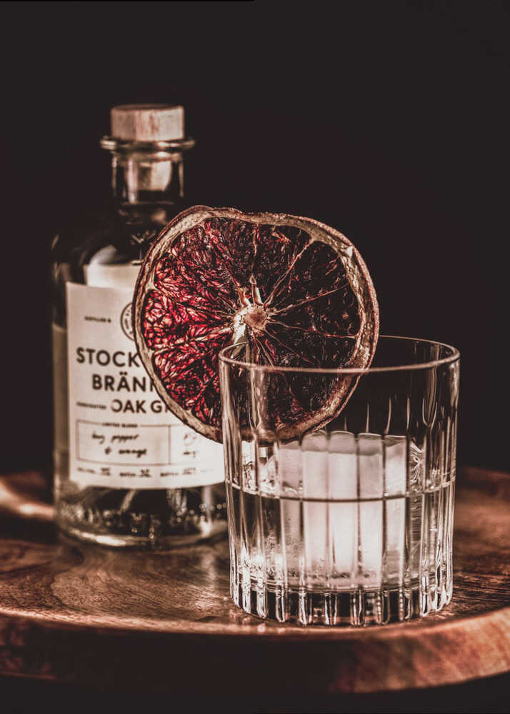 A moody still life photograph of a cocktail in a cut-crystal glass filled with ice. A dried blood orange slice garnishes the rim. In the soft-focus background, a bottle of gin rests on a dark wooden tray against a black backdrop. Wall Art