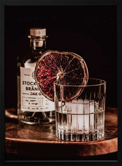 A moody still life photograph of a cocktail in a cut-crystal glass filled with ice. A dried blood orange slice garnishes the rim. In the soft-focus background, a bottle of gin rests on a dark wooden tray against a black backdrop. Wall Art