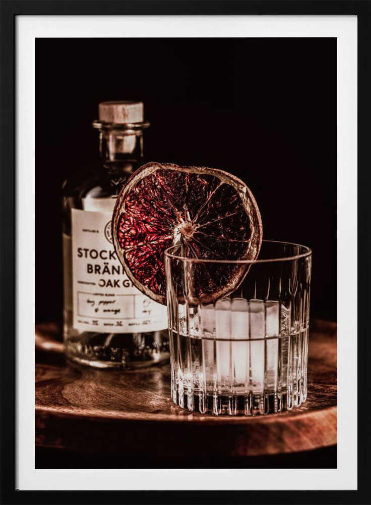 A moody still life photograph of a cocktail in a cut-crystal glass filled with ice. A dried blood orange slice garnishes the rim. In the soft-focus background, a bottle of gin rests on a dark wooden tray against a black backdrop. Wall Art