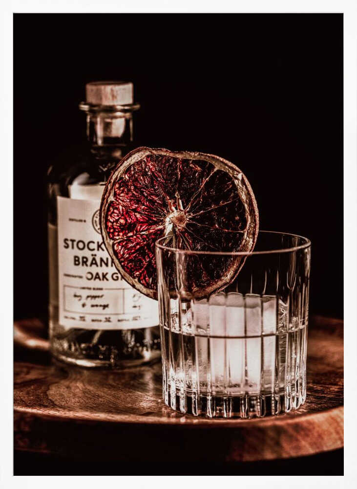 A moody still life photograph of a cocktail in a cut-crystal glass filled with ice. A dried blood orange slice garnishes the rim. In the soft-focus background, a bottle of gin rests on a dark wooden tray against a black backdrop. Wall Art