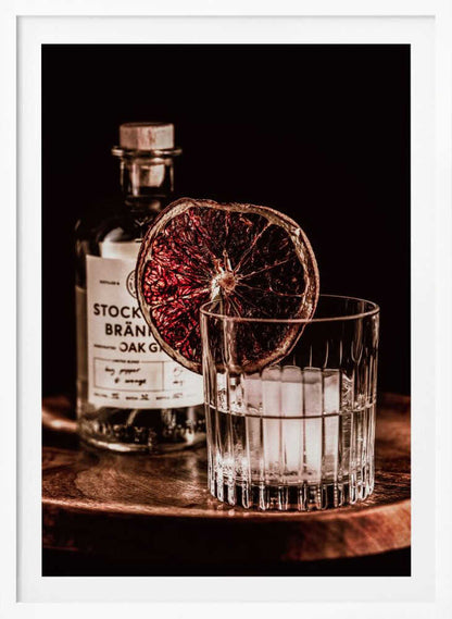 A moody still life photograph of a cocktail in a cut-crystal glass filled with ice. A dried blood orange slice garnishes the rim. In the soft-focus background, a bottle of gin rests on a dark wooden tray against a black backdrop. Wall Art