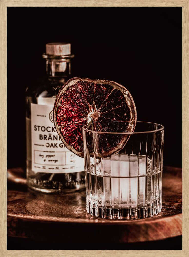 A moody still life photograph of a cocktail in a cut-crystal glass filled with ice. A dried blood orange slice garnishes the rim. In the soft-focus background, a bottle of gin rests on a dark wooden tray against a black backdrop. Wall Art