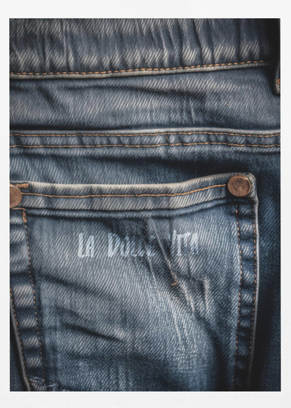 A detailed close-up photograph of a faded blue jean back pocket, showing the texture of the denim, orange stitching, and a copper rivet. Faint white text is stenciled onto the pocket. Poster