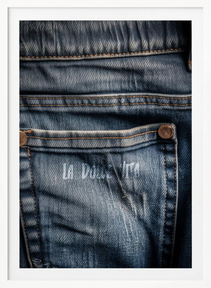 A detailed close-up photograph of a faded blue jean back pocket, showing the texture of the denim, orange stitching, and a copper rivet. Faint white text is stenciled onto the pocket. Poster