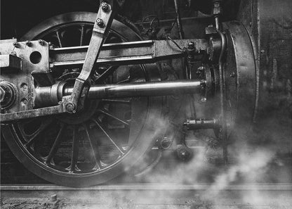 A dramatic, black and white close-up photograph of the wheels and driving rods of a steam locomotive. Puffs of steam rise from the tracks, partially obscuring the lower part of the machinery. Print